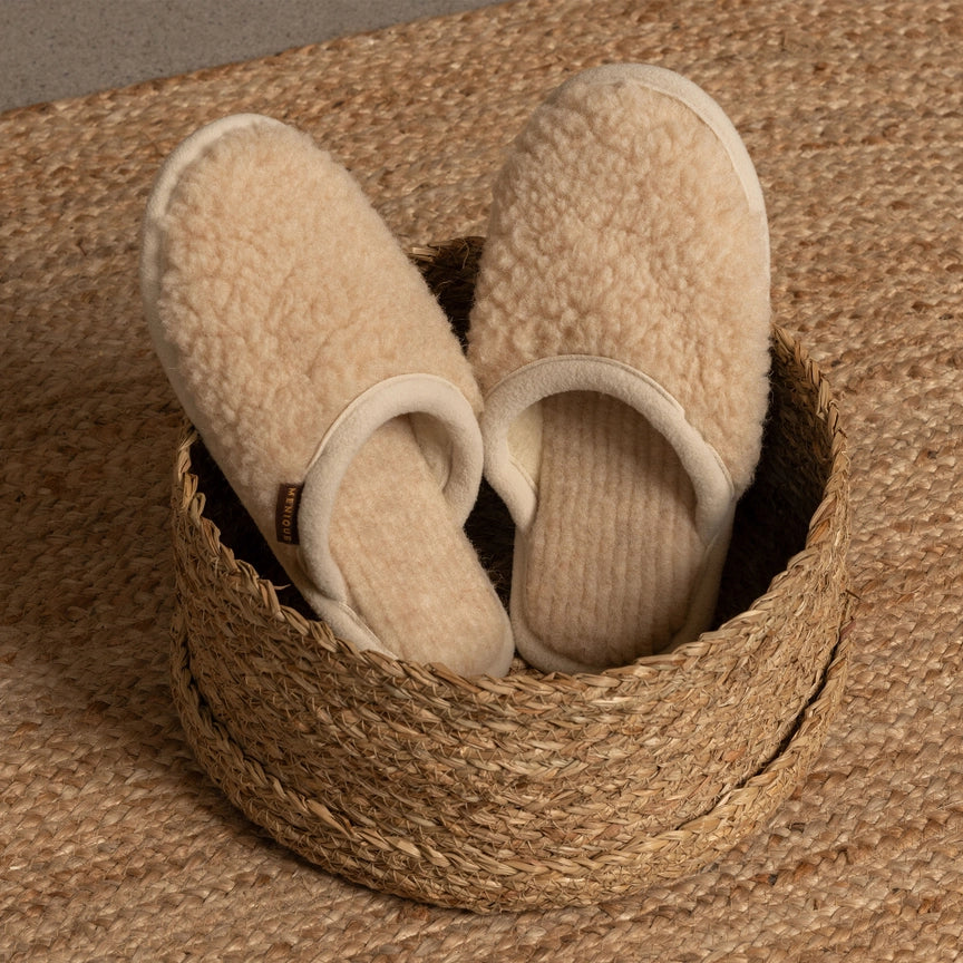 A pair of tan colored eco friendly slipper sit in a wicker basket on the floor. 