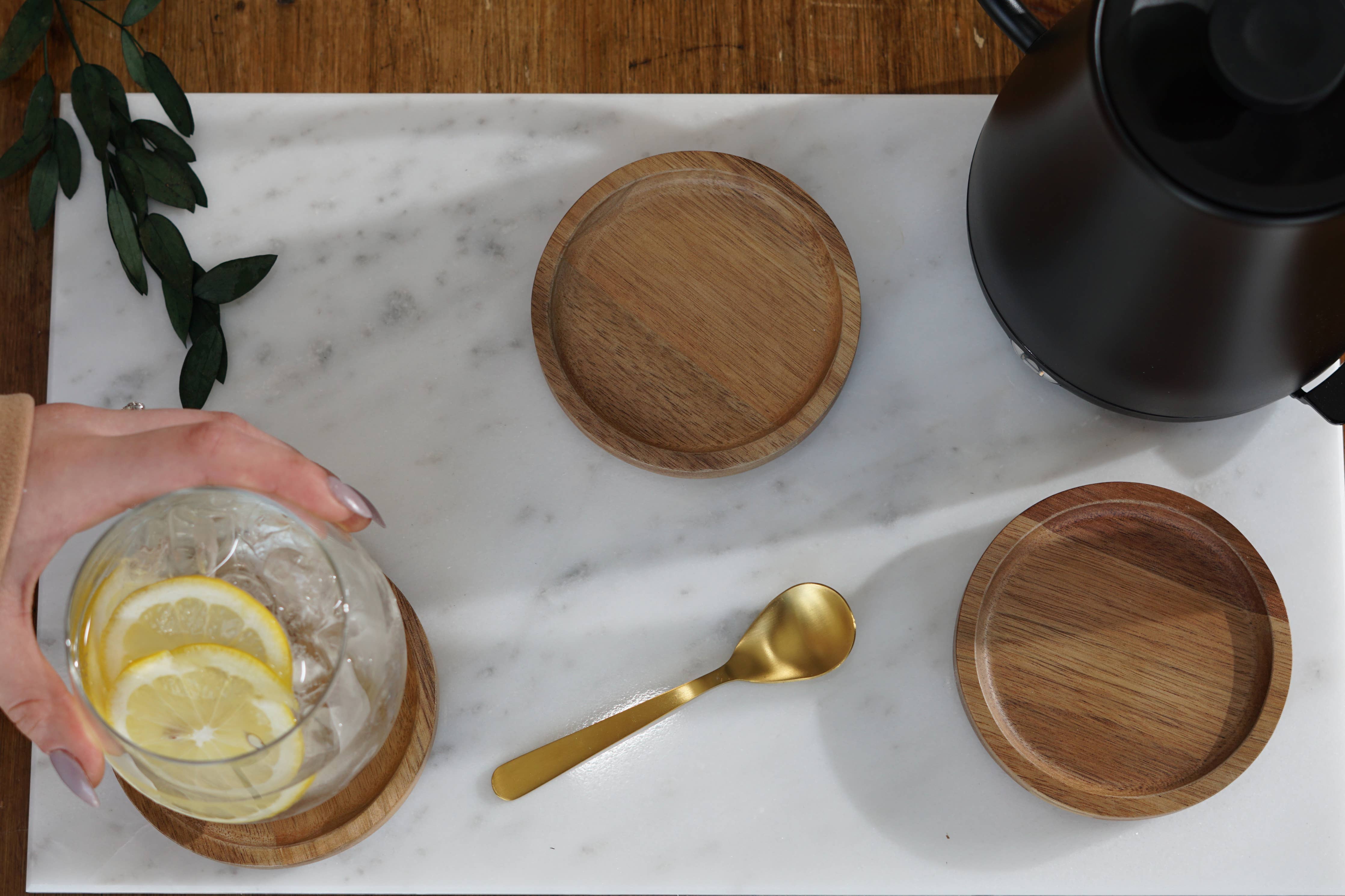 A marble cutting board sits on top of a wooden table. On the cutting board is a coffee carafe, water glass, golden spoon and some wooden coasters. The linked collection contains all of the home and kitchenware products offered by The Good Planet Company. 