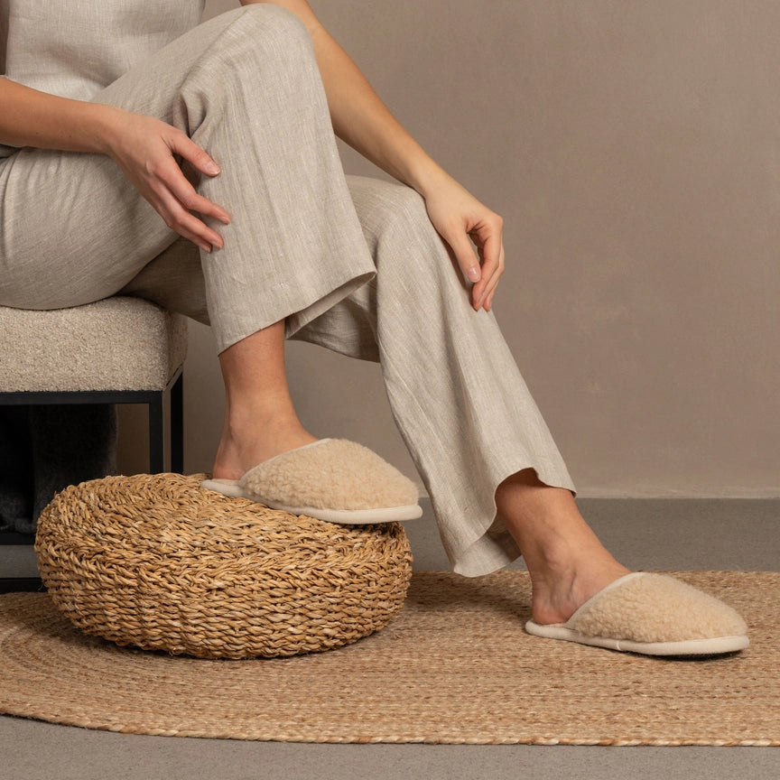 A person sitting on a chair has their foot up on a foot stool. They are wearing eco-friendly slippers on their feet. 