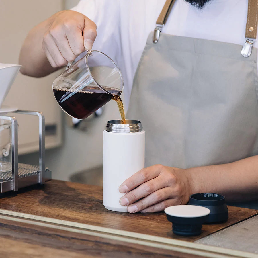 A person pours coffee from a clear glass jug into a white stainless steel travel tumbler. The linked webpage the sustainability blog for The Good Planet Company. 