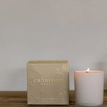 A white glass candle made by Crowfoot Collective sits on a table to the right of the box it was packaged in. 